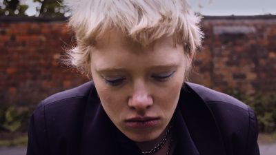 Film still from 'The Path' (2025) by Jon Beaton. Close-up of a blonde, short-haired woman in her late 20s, looking downcast. Behind her a red-brick wall and grey skies.